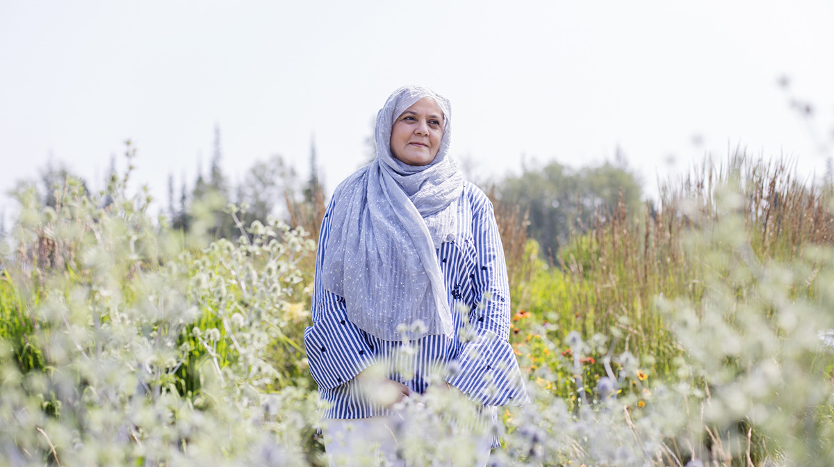 A woman, wearing a long headscarf and striped shirt, stands in the middle of a field of tall wildflowers.